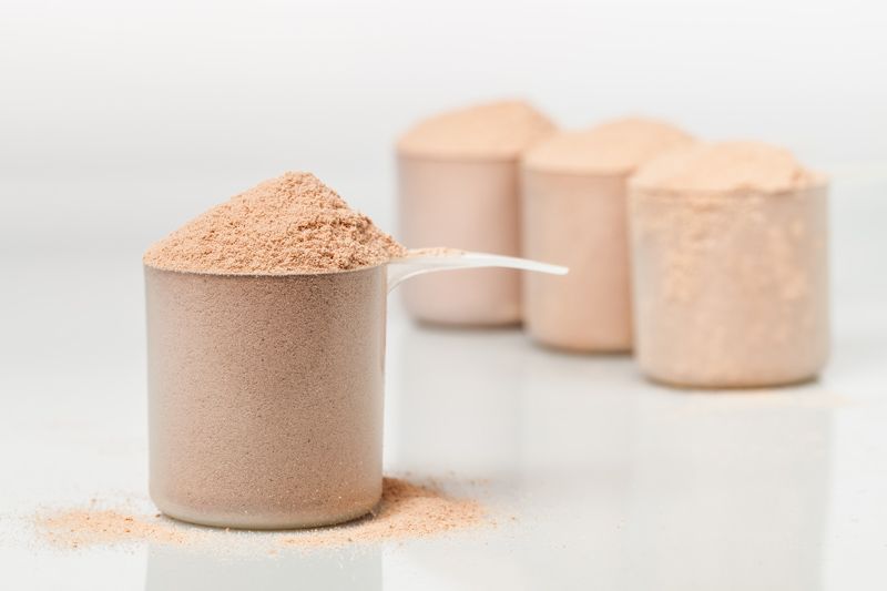 Ein Löffel Whey Protein Isolat Schokolade ohne Aspartam mit drei Löffeln im Hintergrund